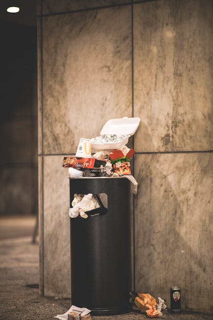 A black cylindrical public waste bin positioned against a beige stone wall in an indoor or covered area, with overflowing takeaway food containers, disposable packaging, and crumpled wrappers stacked on top and spilling out. Additional litter, including a crumpled paper bag and a can, are discarded on the floor nearby. The scene highlights improper waste disposal and cleanliness concerns, often encountered during home relocation or moving processes when removing unwanted refuse as part of packing or clearance, a task that companies like Man With a Van Mayfair assist with. The lighting is neutral, providing clear visibility of the clutter, emphasizing the importance of proper waste management during house removals or moving services in central London.