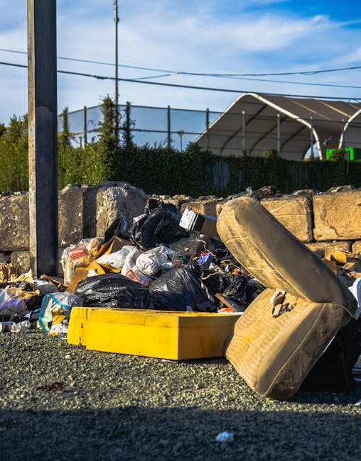 A pile of household waste and discarded items on the side of a street, including black garbage bags, a yellow plastic container, a worn-out upholstered armchair, and various loose packaging materials, situated adjacent to a stone wall with a metal fence, under a clear blue sky with overhead power lines. The waste is on a gravel surface near a pavement, with a utility pole visible on the left. This scene represents improper disposal of bulky waste, highlighting the importance of professional removal services like those offered by Man With a Van Mayfair for house relocations and furniture transport to avoid fines for illegal waste disposal.