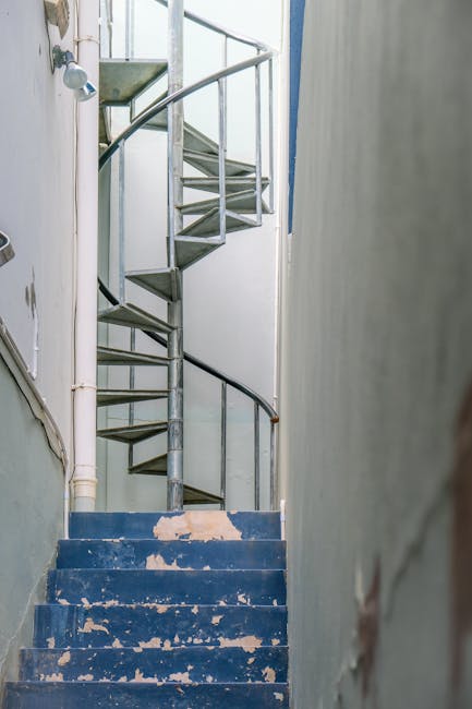 Indoor scene of a narrow, utilitarian staircase with exposed metal steps and a dark wooden handrail, leading upward in a residential property. The staircase is situated between plain, unadorned light-colored walls, with visible pipes running along the left side and an electrical fixture near the top. The steps are painted in a blue hue, though the paint is chipped and peeling, revealing the underlying material. Natural light filters in from a source above, illuminating the stairwell. In the context of house removals, this image illustrates the challenges of navigating tight or awkward staircases during a home relocation. Occasionally, [COMPANY_NAME], such as Man With a Van Mayfair, assists with furniture transport and packing through difficult interior spaces like narrow staircases, ensuring safe loading and unloading of items within properties like this one, aligning with their specialist removal services across Mayfair and Berkeley Square.