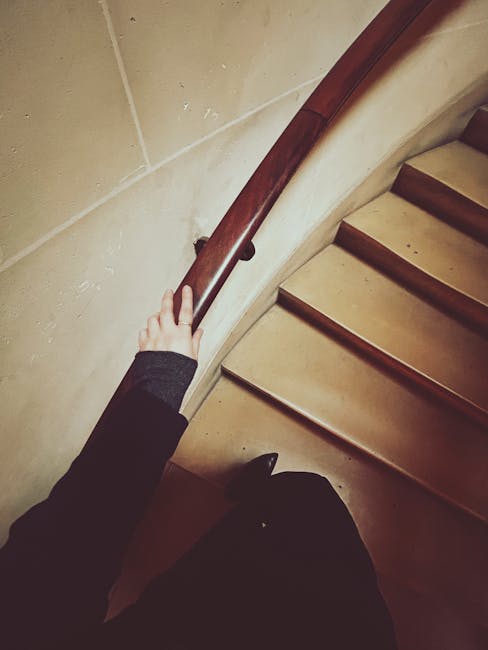 A person wearing a dark long-sleeve top is gripping a wooden handrail on the curved staircase inside a residential property. The staircase has light-colored wooden steps, with a beige or cream wall on the side. The handrail is polished dark brown wood, mounted on small brackets attached to the wall. The environment appears to be well-lit, and the image captures part of the person's arm and hand as they support themselves during a home relocation process. Behind the staircase, there are cardboard boxes and packing materials visible on the floor, indicating an ongoing furniture transport or packing and moving activity. This scene showcases the careful handling involved in house removals, with a focus on navigating narrow staircases when relocating items, as handled by Man With a Van Mayfair from their services in London.