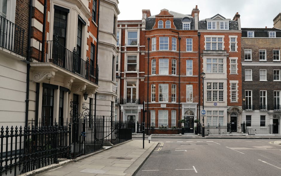 A street scene in Mayfair featuring a row of multi-storey residential buildings with various architectural styles. The building on the far left has a cream-colored facade with black wrought iron balcony railings on the first-floor windows, wooden window frames, and ornate decorative brackets supporting the small balcony. Adjacent to it are taller, red-brick structures with large sash windows, some with white trim, and decorative brickwork around the windows and roofline. The street is paved with grey concrete slabs, with a black metal fence running along the front of the buildings, enclosing small front yards with planters and greenery. In front of the buildings, there is a paved sidewalk, street lamps, and a few traffic signs. The scene is illuminated by natural daylight, indicating a clear or partly cloudy day. The overall setting suggests an area suitable for high-end home relocation, potentially involving furniture transport and packing and moving services, as coordinated by companies like Man With a Van Mayfair.