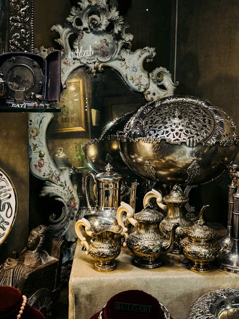 Inside a property during a house removal process, a collection of antique silver and brassware including teapots, urns, and a large decorative bowl with intricate patterns are displayed on a table covered with a beige cloth. Behind the items, an ornate mirror with a detailed white and gold frame reflects part of the room's interior, which features dark walls and framed artwork. To the left, a small shelf holds a decorative plate and other objects. The setting suggests preparation for packing and furniture transport, with the visible objects carefully arranged for a move. This scene exemplifies the types of antique items handled during a specialist relocation service provided by Man With a Van Mayfair, as part of their removals and home relocation offerings. The lighting highlights the polished surfaces of the antique metalware and the detailed craftsmanship of the mirror.