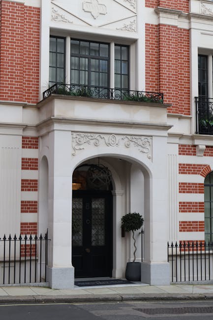 The image shows the exterior of a multi-storey building with a classic architectural style, featuring a facade constructed from red brick and white decorative stone accents around windows and doorways. The ground floor has an arched entranceway with a black door, flanked by two large windows with ornate iron railings on the balcony above. The entrance is framed by a white stone arch with decorative carvings, and a small potted tree is placed on each side of the door, positioned on the pavement. The building's upper floors have additional windows with white trim, and the facade includes detailed stonework and decorative elements. A black metal fence runs along the sidewalk in front of the building. The scene suggests a residential or commercial property in an urban setting, typical of a Mayfair address, and the context alludes to professional house removals or furniture transport services, with no moving activity directly visible in the image.