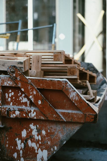 A close-up view of a rusty, weathered boat with white paint splatters along its surface, docked near a modern residential building with large glass windows and white framework. Inside the boat, there are neatly stacked wooden pallets and plastic crates, likely used for shipping or moving furniture and boxes. An orange strap is visible, possibly used for securing items during transportation, and the boat appears to be prepared for a home relocation or furniture transport process handled by Man With a Van Mayfair. The scene shows outdoor environment with natural light illuminating the workspace, indicating an ongoing packing and loading operation for a house removal or moving service in Mayfair, with the potential for loading items onto a vehicle for delivery.