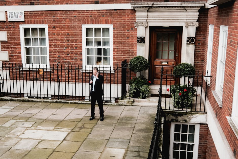 A man in a dark suit stands on the paved pavement outside a red-brick residential building, speaking on a mobile phone. The building features white-framed windows with sheer curtains, a dark wooden front door set within a decorative stone surround, and small potted plants with green foliage and red flowers positioned on either side of the entrance. A black metal fence encloses the small front yard area. Visible in the image is a white sign displaying the street name 'Grosvenor Square W1'. The scene captures the exterior environment during daylight, with even lighting highlighting the building's architecture and surrounding details, relating to home relocation or furniture transport services provided by Man With a Van Mayfair, as part of professional removals or moving logistics.