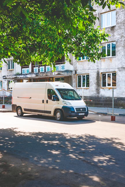 A white cargo van operated by Man With a Van Mayfair is parked on a paved street in front of a residential building, with large leafy green trees providing partial shade. The van is positioned parallel to the sidewalk, near a curb, and appears to be in the process of a home relocation or furniture transport, indicative of professional removals services. Behind the van, the building features multiple rectangular windows, some with external decorations, and shows signs of weathered concrete exterior walls. The scene suggests a quiet urban environment suitable for house removals or moving logistics, with the van ready for loading or unloading furniture and boxes, possibly in the process of packing or preparing for transport. Equipment such as blankets, plastic wrap, or trolleys is not visible, but the setting aligns with typical moving service activities managed by Man With a Van Mayfair.