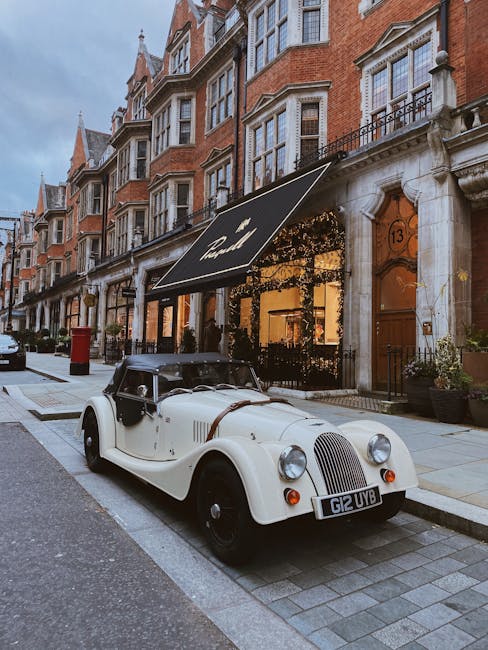 A vintage white open-top car with black accents is parked on a cobblestone street outside a Georgian-style building with large sash windows and ornate brickwork in Mayfair. The car's registration plate reads 'G12 UVB'. The building features a black awning with the logo of a hospitality or retail venue, decorated with string lights and greenery, suggesting a commercial property. Potted plants are positioned beside the entrance, and other parked vehicles can be seen further down the street. The scene is set during daylight with soft, natural lighting, and the street appears calm and clean, ideal for home relocation or furniture transport. As part of a house removal or moving process, [COMPANY_NAME] might be involved in managing or coordinating vehicle access and logistics in this area.