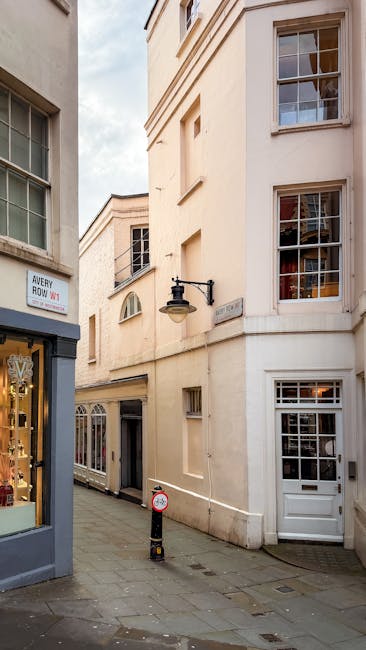 A narrow street corner in a residential area with cream-colored, multi-storey buildings featuring large sash windows and decorative architectural details. On the left, a small shop with a display window and a sign reading 'Avery Howe' is visible, while on the right, a white door with a glass panel provides access to a property. The street is paved with grey cobblestones and a black bollard is positioned near the curb. An outdoor wall-mounted lamp is attached to one of the buildings, illuminating the area. The scene depicts a typical street scene in Mayfair, London, with no visible moving activity but serves as a suitable background for house removals and furniture transport themes, illustrating the environment where professional house removals services such as those offered by Man With a Van Mayfair might operate during home relocation or packing and moving processes.
