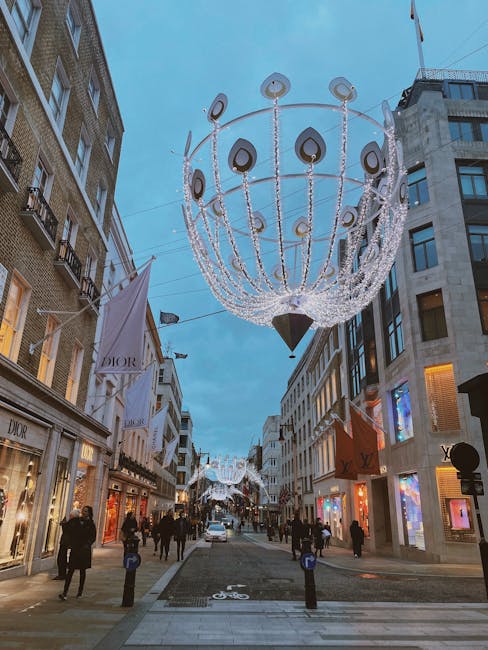 A daytime street scene in Mayfair decorated with large, illuminated festive chandeliers hanging above. The chandeliers are spherical, composed of multiple strands of white lights and black teardrop-shaped accents, suspended from wires between tall buildings on either side of the street. The storefronts on the left include Dior and other luxury brands, with large display windows and branded signage. Pedestrians are walking along the pavement, some carrying shopping bags, while a few are stationary, possibly observing the decorations or engaged in conversation. At the intersection ahead, a white van is parked, and the street features bicycle lanes marked on the paved surface. The surrounding buildings are modern and stylish, with large windows and neutral-toned facades. The lighting suggests early evening or late afternoon, with a clear sky overhead. The scene captures an active shopping district with seasonal or event-based decorations, and [COMPANY_NAME] provides professional assistance with moving or logistical services in the area.