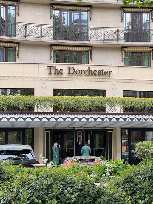 The exterior of The Dorchester hotel in Mayfair, London, features a cream-colored facade with multiple large windows and black iron balcony railings on the upper floors. A green hedge runs along the front of the building, partially obscuring the ground-floor entrance. The entrance has a black canopy with decorative white trim, and two staff members in green uniforms are seen near the doorway, engaged in a task related to a home relocation. In front of the hotel, a black van is parked on the street, ready for furniture transport or packing and moving activities. The scene suggests a professional relocation process possibly coordinated by Man With a Van Mayfair, focusing on boutique office or residence moves with minimal downtime, within a setting that emphasizes precision and professionalism in logistics and furniture transport.