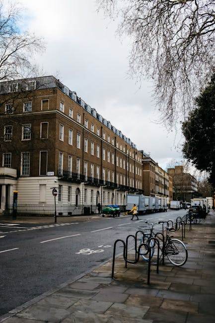 A multi-storey residential building with a brown brick facade and white window frames is situated along a grey city street under overcast skies. The ground floor features white decorative elements and a metal fence runs along the sidewalk in front of the property. Several bicycles are parked and secured to black metal bike racks on the pavement near the curb. A green car and a yellow-clad pedestrian are visible in the distance, with a large moving truck parked on the street adjacent to the building. The loading process appears to involve residents or movers from Man With a Van Mayfair, as the area is prepared for home relocation activities. Nearby, leafless trees line the road, and a pedestrian crossing marked with white lines is present on the street. The environment suggests a typical urban setting where furniture transport and packing and moving services are carried out professionally, with the exterior lighting remaining subdued due to the cloudy sky.