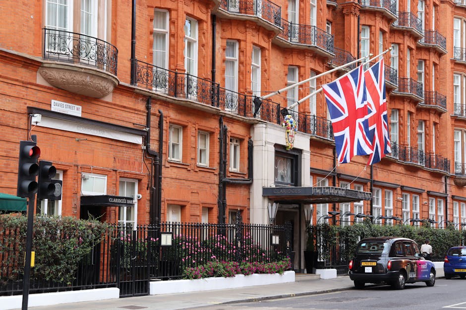 A multi-storey red brick building on a city street, featuring several black wrought iron balconies with decorative railings and large sash windows. A Union Jack flag is draped across the facade, attached to cables strung between the building and poles. In front of the building, a black metal fence surrounds a small garden with pink flowers and green shrubs. A street sign indicates the location as Davies Street W1. On the pavement, a man is seen carrying a large cardboard box, while nearby a black taxi and a blue car are parked. To the left, a traffic light is visible, and an entrance to a property named 'Claridge’s Bar' can be seen beneath an awning. The scene suggests a busy urban environment with activity related to house relocation or moving services, as coordinated by companies like Man With a Van Mayfair, with a focus on logistics involving packing, furniture transport, and loading processes.
