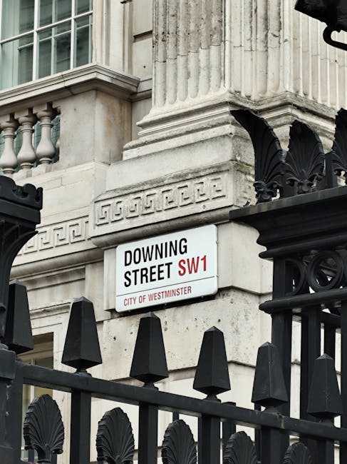 The image shows a section of a historic building with intricate stonework and decorative columns, partially obscured by a black wrought iron fence featuring decorative finials and floral motifs. Attached to the building is a street sign indicating 'Downing Street SW1' under the jurisdiction of the City of Westminster. In the foreground, a man wearing a dark jacket and gloves is seen lifting or carrying a large, wrapped piece of furniture covered with protective fabric and plastic wrap, possibly part of a home relocation or furniture transport process. The scene is set outside, with natural daylight illuminating the stone facade and the surrounding environment, illustrating a professional moving or packing operation coordinated by Man With a Van Mayfair, specializing in removals and house relocation services.